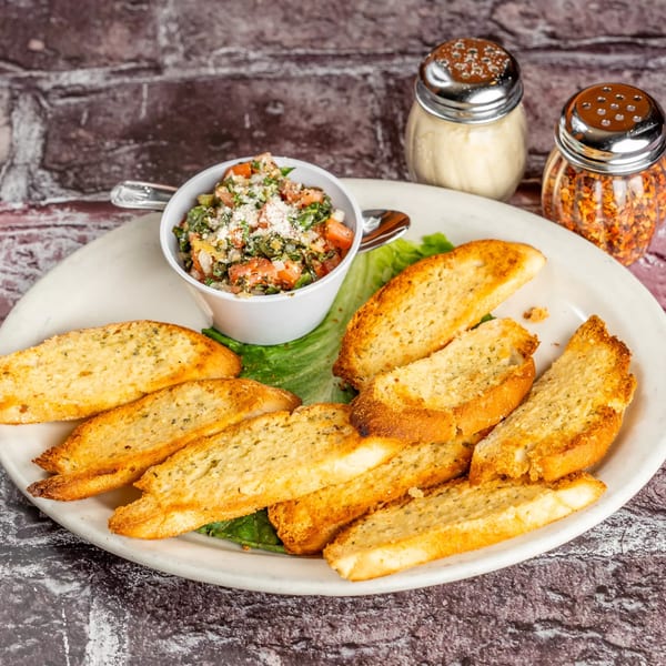 A plate of toasted garlic bread slices with a side of bruschetta, Parmesan, and red pepper flakes.